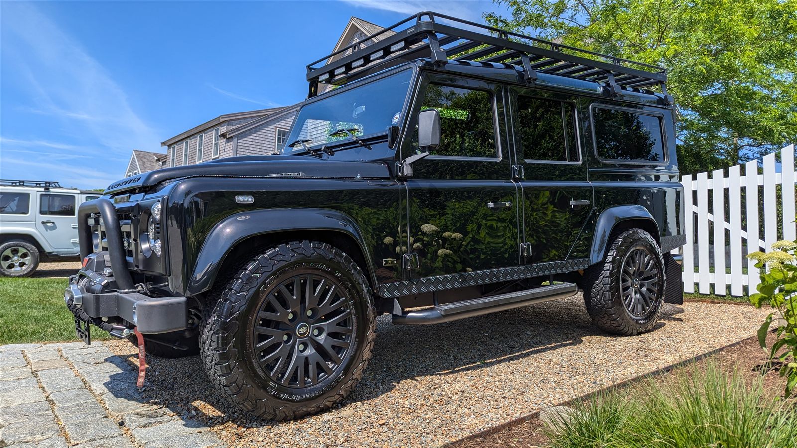 Black Land Rover Defender with deep mirror-gloss paint reflecting hydrangeas, beside a white picket fence